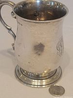 Front angled view of the silver cup showing the body shape, handle and hallmarks near the rim alongside a coin for scale.
