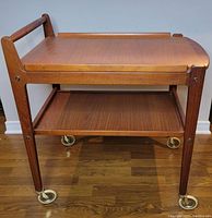 Front three-quarter view of assembled teak bar cart showing two tiers, legs, castors and removable top tray