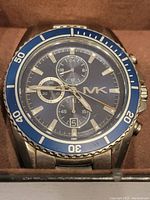 Close-up of the watch face showing 'MK' logo, blue bezel with minute markers, and chronograph subdials.