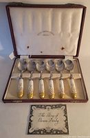 Original fitted case open to show six Royal Crown Derby spoons inside with ceramic handles and gold accents, plus informational booklet.