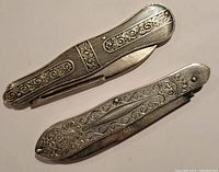 Closed view of two silver-plated vintage pen knives showing intricate raised scroll and floral patterns on the handles.