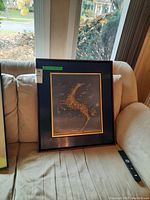 Framed artwork featuring a horse formed of golden leaves, positioned on a couch with window light in the background. Shows minor scratches on the frame.