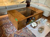 Front and side angle showing the wood coffee table with glass center top and two compartments below, on a patterned rug with couches in background.