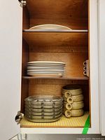 Wood cabinet shelf with 12 stacked white ceramic plates, one large white platter above them, six clear glass sectional plates stacked, and six speckled ceramic soup bowls with handles stacked.