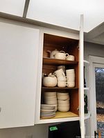 White ceramic oven-to-tableware set including teapot and stacked bowls and cups in kitchen cabinet.