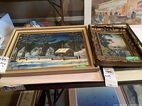 Two paintings side by side on a table: one larger winter scene by A.M.M. Watson, one smaller ornate framed landscape by Georgie Wilcox.