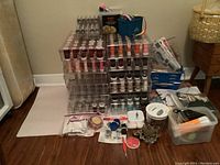 Overview of 12 acrylic storage bins stacked, containing multiple spools of thread in various colors, along with multiple packages and small containers of sewing accessories including pins, bobbins, and tools.