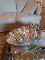 Full view of silver tea service on footed tray with teapot, coffee pot, creamer and sugar bowl arranged on a cloth, background showing a sofa.