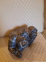 Soapstone figurine displayed on a beige textured chair showing the side profile of the figure in a reclining pose.