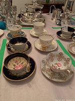 Photo showing six bone china tea cups and saucers in various patterns, including floral and gold designs, on a table with other silverware in the background.