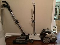 Side view of silver Hoover vacuum cleaner with WindTunnel technology, large wheels, and attached hose and metal wand, alongside floor brush and upholstery head attachments leaning against wall.