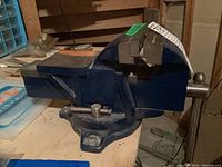 Side view of blue metal bench vice showing adjustable jaws, screw handle, and swivel base mount.