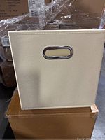 Side view of beige cloth bin showing metal oval handle and fabric texture.