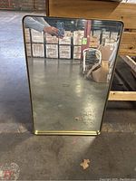 Front view of rectangular wall mirror with gold metal frame, clear glass surface, standing on warehouse floor.