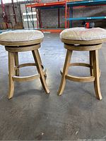 Side view of two swivel bar stools with light wood finish and fabric wrapped seats showing legs and footrest rings