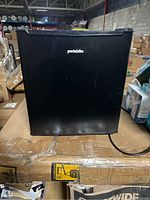 Front view of black Proctor Silex mini fridge with logo, sitting on pallet in warehouse.