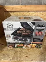 Front view of the Ninja Foodi Smart Grill box highlighting grilling features and images of cooked food.