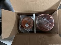 Two terra-cotta planters sealed in plastic inside a cardboard box, showing the planters’ cylindrical shape and warm brown color.