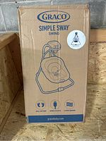 Closed original cardboard box of Graco Simple Sway baby swing in brown with blue printing showing illustration of swing and branding.