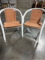 Pair of two wicker chairs shown front view with protective wrapping on white metal frame and light brown woven seats and backs.