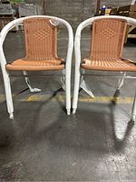 Two wicker chairs with woven tan rattan seats and backs, white metal frames, showing front view on industrial floor.