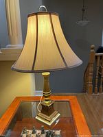 Two photos of one table lamp showing fabric shade with ribbing and slight stains near bottom edge, brass-tone base with stepped square design and ridges.
