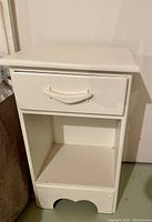 Front angled view of one white small side bed table showing drawer, open shelf, and decorative scalloped cutout at base.
