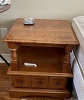 Photo of a solid wood bed side table with three lower drawers and an open shelf between the top and drawers. Table has two turned supports and a smooth flat top.