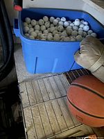 Blue plastic bin filled with numerous used white golf balls showing dirt and wear, placed on a metal rack next to basketball and golf head cover.