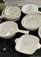 Set of five Corel CorningWare dishes with lids on a black stovetop. White with blue cornflower design. Various shapes and sizes visible.