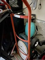 Photo of an orange hand truck dolly holding a blue gas or oxygen tank secured with a chain. The dolly has black and white wheels and a small foot platform.