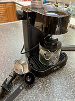 Black Bella electric espresso maker on kitchen counter, with glass carafe positioned on drip tray, portafilter and measuring scoop shown in front.