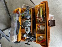 Photo of open orange fishing tackle box displaying compartments filled with fishing lures, lines, and accessories with three fishing reels resting inside.