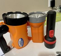 Photo showing three flashlights: large orange/black with handle, medium orange/gray, and small black/red.