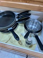 Multiple used frying pans stacked alongside a stainless steel pot with a steamer insert and lids in a kitchen drawer.