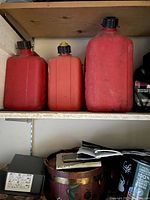 Photo of three plastic gas containers in red and orange on a shelf, showing used condition with dirt and faded surfaces.