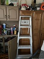 Four-step aluminum ladder leaning against a wooden cabinet in a garage or workshop environment, surrounded by other household items and cleaning products.