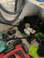 Wide angled photo showing the green lawn mower with black engine, folded handle, white wheels, and some cloths on top, stored indoors.