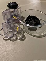 Photo showing multiple clear plastic Magic Bullet cups of various sizes, black lids and caps, and a large glass bowl containing some lids.