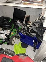Photo of a blue foldable mobile scooter with black cushioned seat and pedals, placed on floor alongside various other items including a lawn mower and bicycle parts.