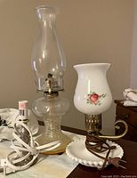 Photo shows two lamps: a vintage clear glass oil lamp with chimney and a white floral electric lamp with frosted glass shade, each with their cords visible.