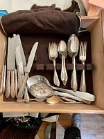 Top view of a drawer with silverware including forks, knives, spoons, serving spoons arranged inside a soft brown cloth-lined drawer.