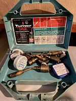 Open green plastic carrying case showing Turner Torch label and several soldering torch tips, solder wire rolls, and a small metal container.