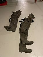 Photo showing two pairs of greenish-brown wader overalls with attached boots laid flat on a light floor, highlighting their full length, boot soles, and adjustable shoulder straps.