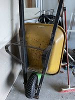 Side angled view of used yellow metal wheelbarrow showing black metal handles and single black pneumatic tire, some rust and wear visible.