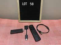 Amazon Firestick 2nd generation device, remote, HDMI extender, and USB power cable displayed on a pink surface with 'Lot 56' sign in background.