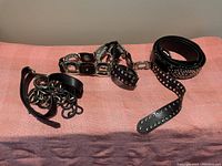 Three women's fashion belts in black faux leather with various silver-tone metal decorative designs, laid on a pink fabric background.