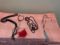 Image of four costume jewelry necklaces and one bracelet displayed on a pink and purple fabric background, showing black beaded tassel necklace, purple bead necklace, silver chain with red feather pendant, silver chain with blue tassel pendant, and thin silver bracelet with dangling chain.