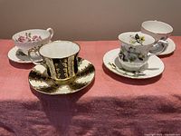 Photo showing four different vintage tea cups and saucers with floral and gold detailed patterns, arranged on a pink cloth.