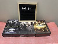 Photo showing all three Deadwood DVD box sets for seasons one to three placed side by side underneath a board labeled LOT 68.